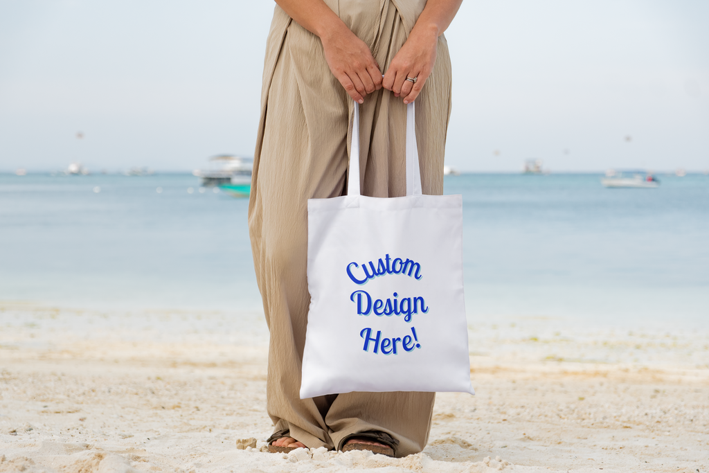 Person holding a white tote bag with 'Custom Design Here!' text on a beach.